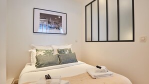Ein ordentlich bezogenes Bett mit weißer Decke und Kissen, ein gerahmtes Bild an der Wand und ein großes Fenster mit Jalousien.