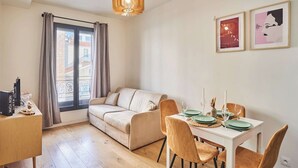 Een moderne woonkamer met een bank, een eettafel gedekt met borden en glazen, een kroonluchter en twee ingelijste kunstwerken aan de muur.