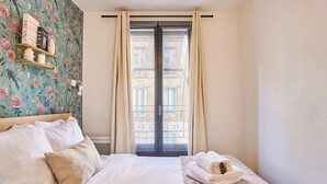 Een slaapkamer met een bed, een nachtkastje met boeken, een raam met gordijnen en een uitzicht op gebouwen.