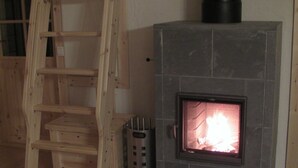 Una chimenea de piedra con fuego encendido, una escalera de madera apoyada en ella y un cenicero de metal al lado de la chimenea.