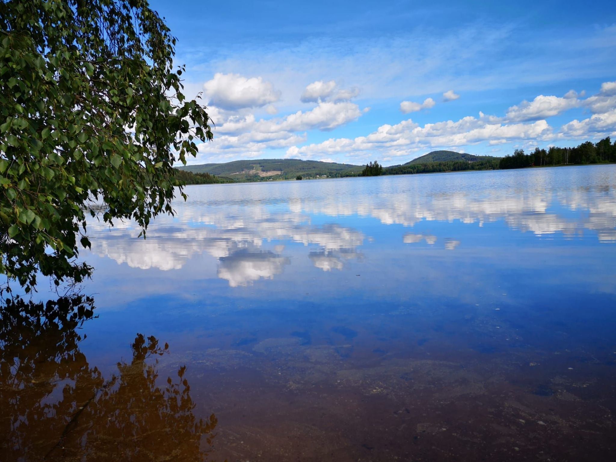 A calm lake with a clear reflection of the sky and clouds, surrounded by hills and trees.