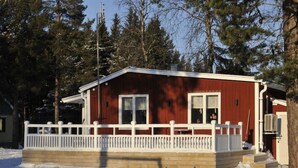 Une cabane rouge avec un porche blanc et une petite terrasse, entourée de neige et d’arbres.