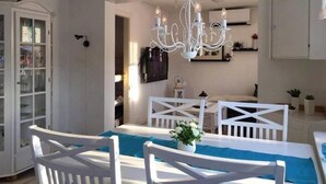 Un coin repas avec une table et des chaises blanches, un lustre, et une vue sur une pièce où se trouve un lit.