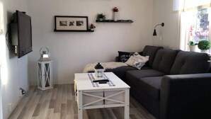 Un salon comprenant un canapé, une petite table blanche, un téléviseur fixé au mur, un climatiseur mural et un tableau encadré.