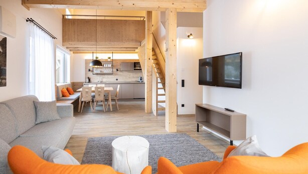 A modern living room with a sofa, orange chairs, a TV, and a staircase leading to a kitchen.