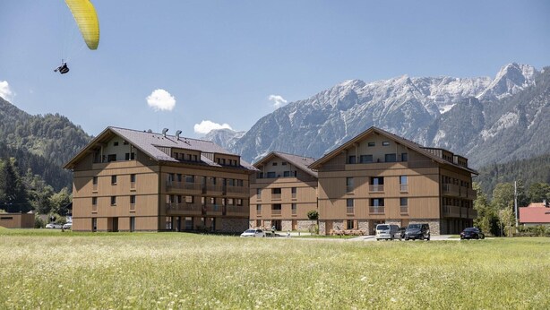 A multi-story building with balconies, a field in the foreground, and mountains in the background.