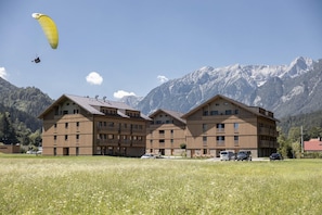 A multi-story building with balconies, a field in the foreground, and mountains in the background.
