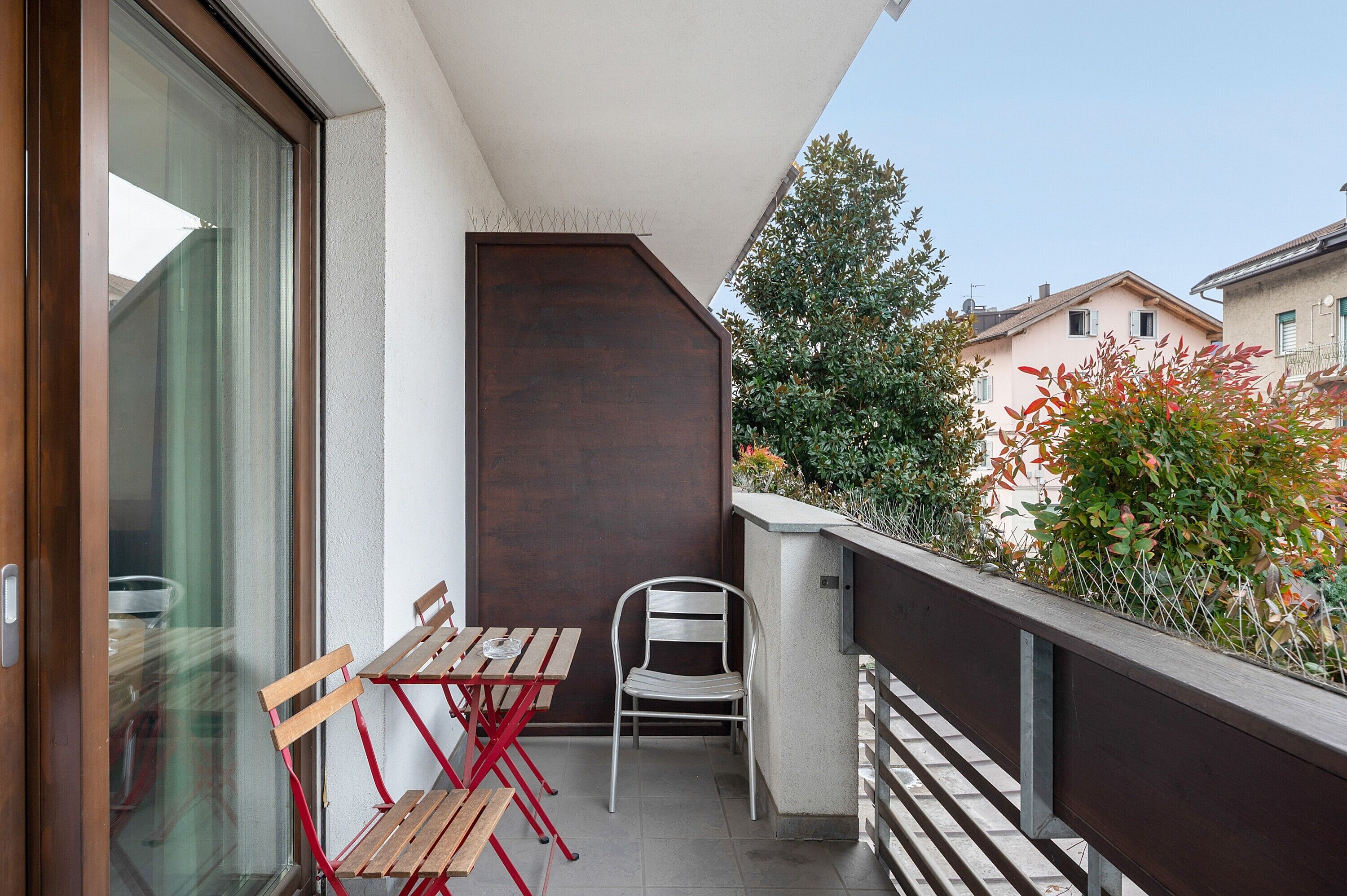 Ein Balkon mit Holztisch und Stühlen, einem Metallstuhl und Blick auf benachbarte Häuser und Grünpflanzen.