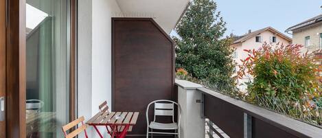 Ein Balkon mit Holztisch und Stühlen, einem Metallstuhl und Blick auf benachbarte Häuser und Grünpflanzen.