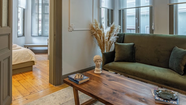 Une pièce avec un canapé vert, une table basse en bois et un buste décoratif posé sur la table. Une porte donne accès à une autre pièce où se trouve un lit.