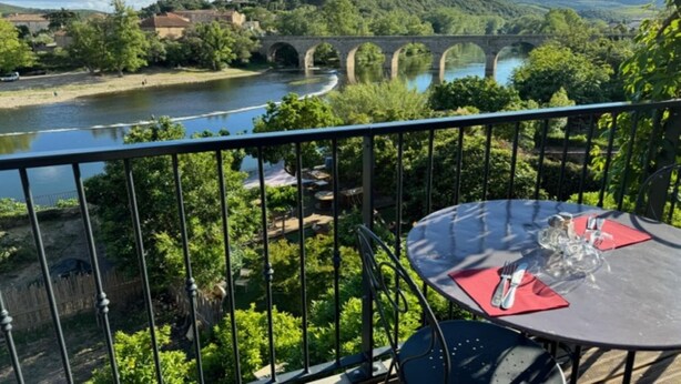 Un balcon avec une table dressée pour le dîner, donnant sur une rivière et un pont.