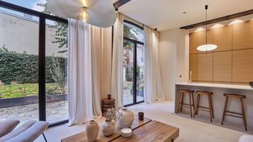 A modern living room with a large window, a wooden table with vases, and a kitchen area with stools.