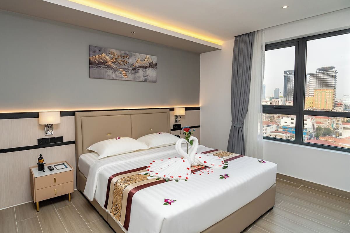 Habitación de hotel con una cama grande, una mesita de noche, una lámpara de pared y vistas a la ciudad a través de un ventanal amplio.