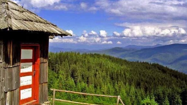 Uma cabana de madeira com porta vermelha, cercada por uma vedação de madeira, emoldurada por uma floresta densa e montanhas sob um céu parcialmente nublado.