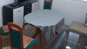 Ein Essbereich mit einem runden Tisch, zwei Stühlen, einer Couch, einem Fernseher auf einem Schrank und einem Fenster mit Jalousien.