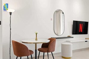 A modern hotel room with a round dining table, two brown upholstered chairs, a white vase with flowers, a floor lamp, a large mirror, a TV with a Netflix logo, and a white trash bin. - The Velvet Apex Studio w/ Jacuzzi Trillionaire (Dubai)