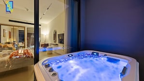Een moderne hotelkamer met een ruime, verlichte jacuzzi, een glazen wand met uitzicht op het interieur, en een netjes opgemaakt bed met handdoeken.