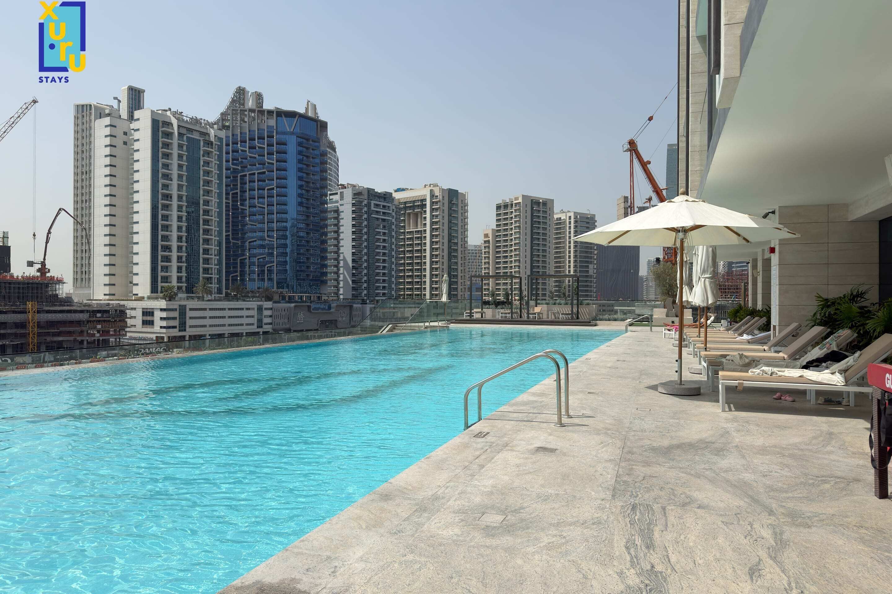 A swimming pool with a cityscape in the background, lounge chairs, and a construction crane.