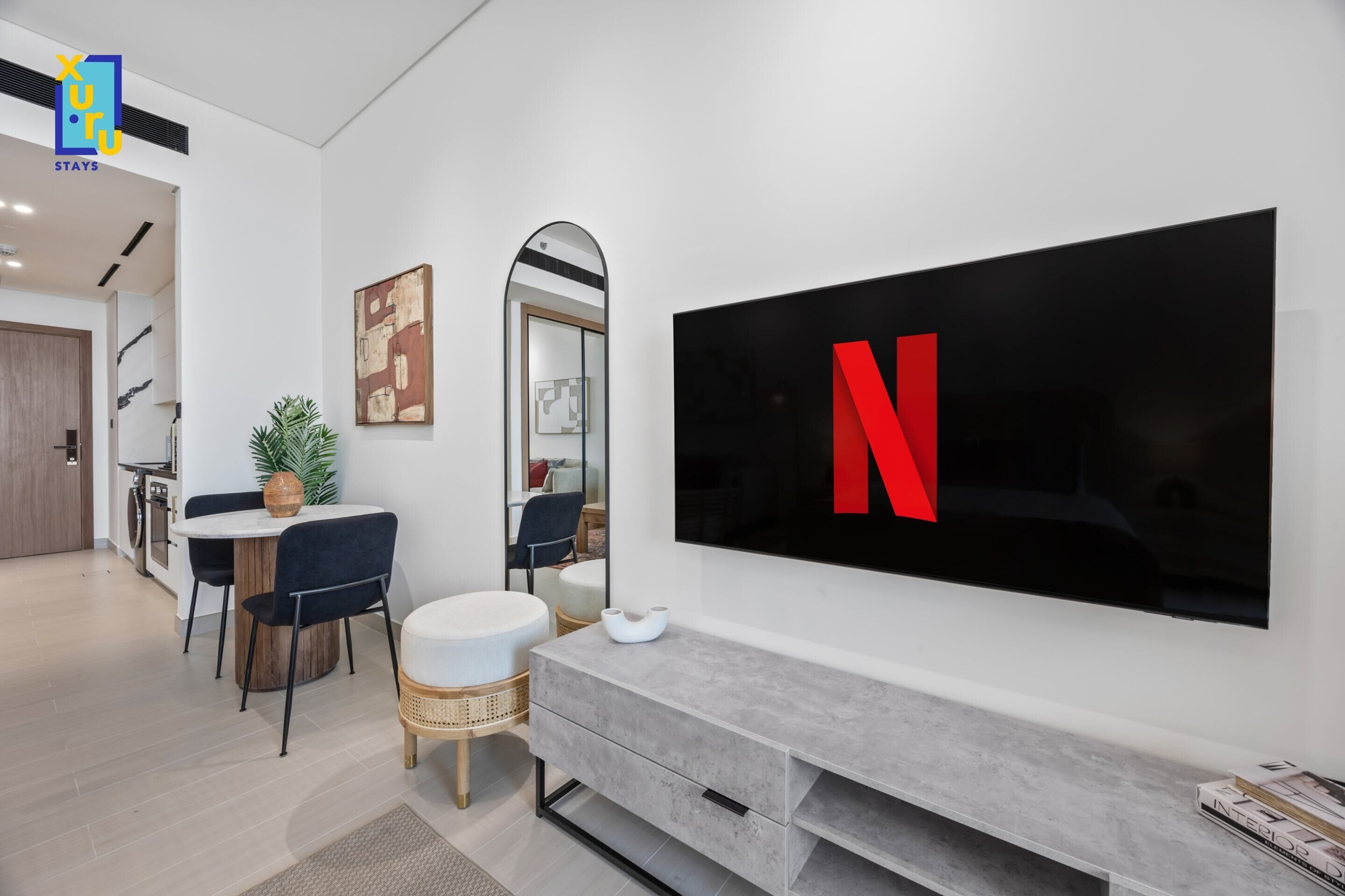 A modern living room with a large flat-screen TV mounted on the wall, a dining area with a round table and chairs, and a kitchen visible in the background.