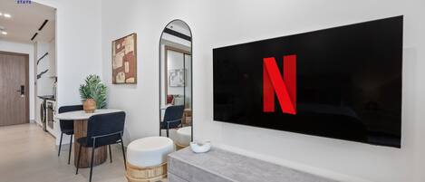 A modern living room with a large flat-screen TV mounted on the wall, a dining area with a round table and chairs, and a kitchen visible in the background.