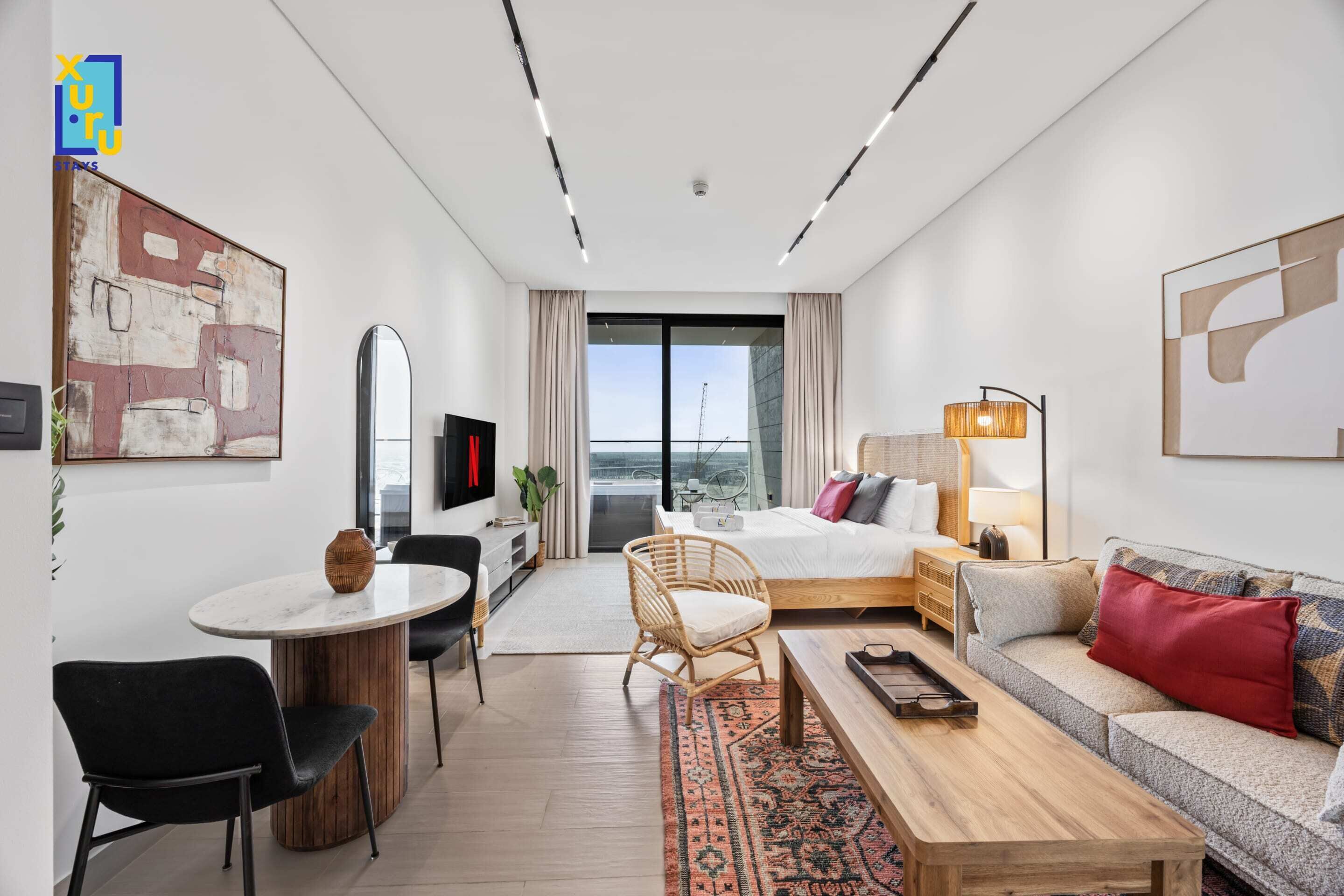 A modern hotel room with a bed, sofa, dining area, and a large window with a view.