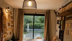 A dining area with a long wooden table, white chairs, a hanging light fixture, and a view through a glass door to a garden. - Domaine du Pin – Nature Escape for Groups (15)+ Spa & Activities -South Brittany (LANGUIDIC)