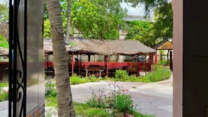 An outdoor seating area with a thatched roof, surrounded by greenery and a paved walkway.