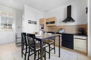 Une cuisine moderne avec une table à manger, des chaises, des placards, un four noir et une fenêtre surmontée d’un auvent à rayures.