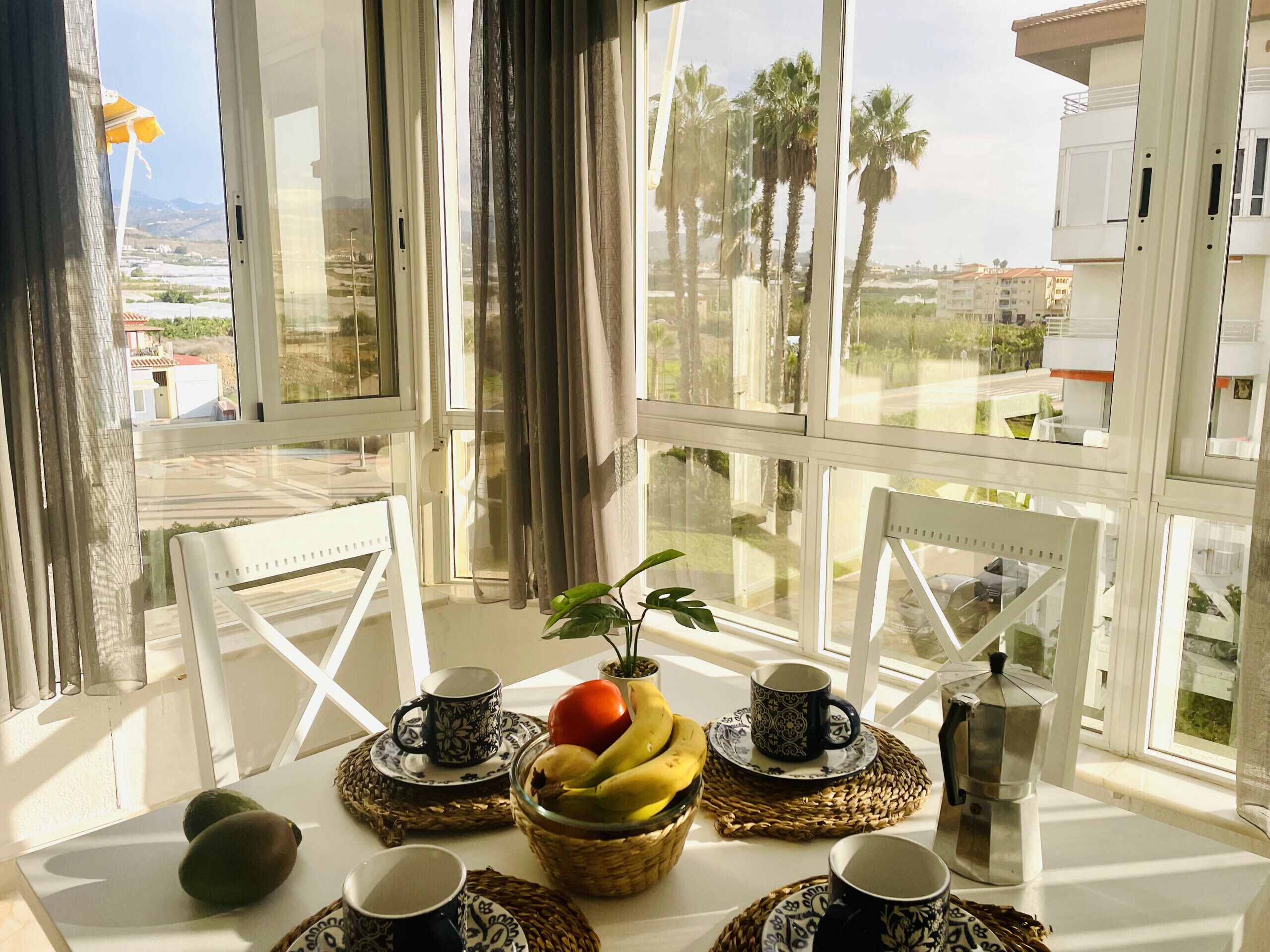 A table with a fruit bowl, two cups, and a coffee pot, set against a window with a view of palm trees and buildings.