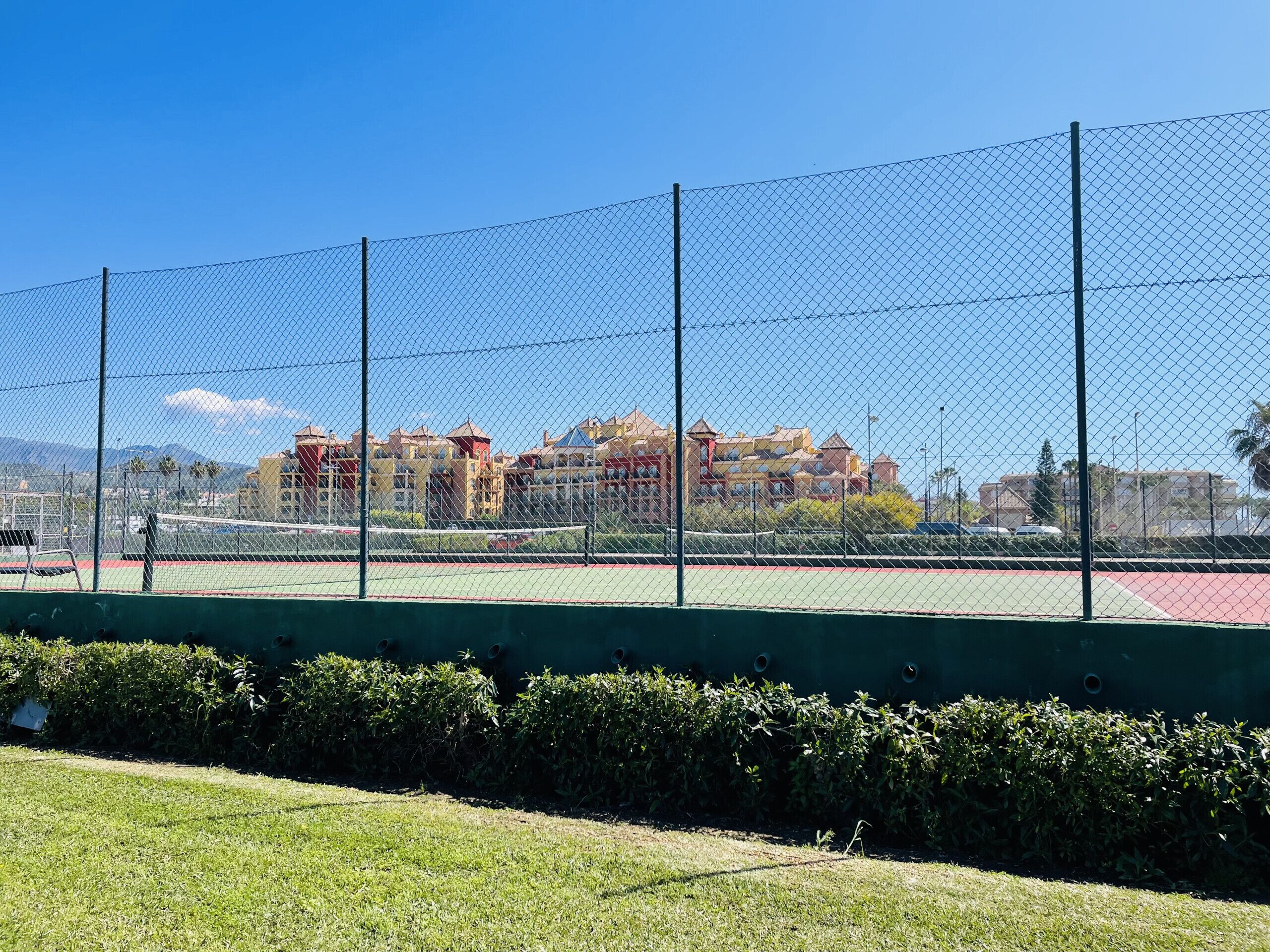 En tennisbana med staket och grön yta, omgiven av en häck och en rad byggnader i bakgrunden.
