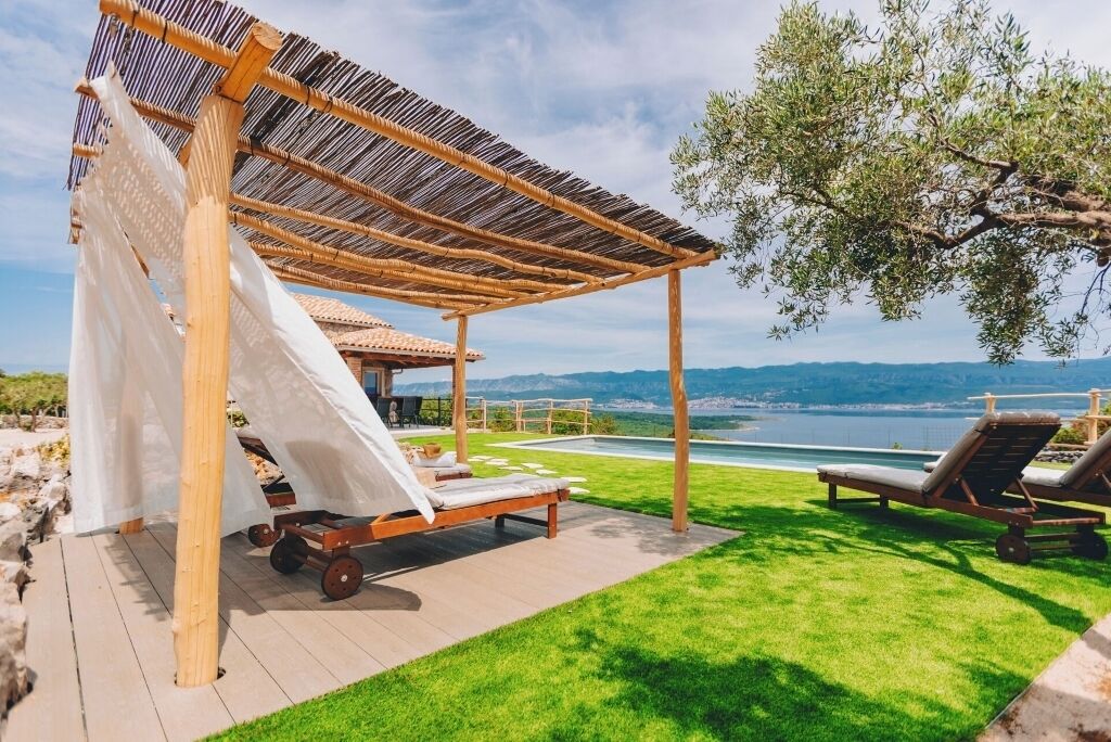 En pergola av trä med ett vitt tak, två solstolar och utsikt över en sjö eller ett annat vattendrag samt berg.