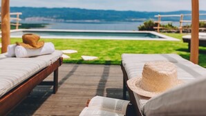 Een houten terras met twee ligstoelen, waarvan er een met een strohoed is voorzien, met uitzicht op een zwembad en een prachtig panorama van bergen en een waterpartij.