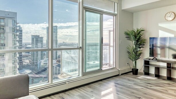 A modern apartment with large floor-to-ceiling windows, a flat-screen TV, and a potted plant. - DT Views | UG Park | Walk to Saddledome (Calgary)