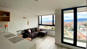 A modern living room with a large window, a sofa, a coffee table, and a dining area with a white table and chairs. - Penthouse in Zona T with Rooftop Pool, Sauna & Gym (Bogotá)