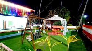 Un espacio exterior bien iluminado con luces artificiales, un parque infantil con columpios, una zona cubierta con asientos y césped cuidado.