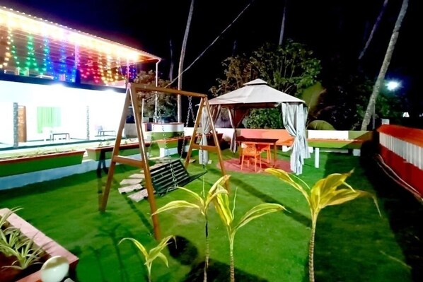 A well-lit outdoor area with artificial lighting, a swing set, a covered seating area, and neatly trimmed grass.