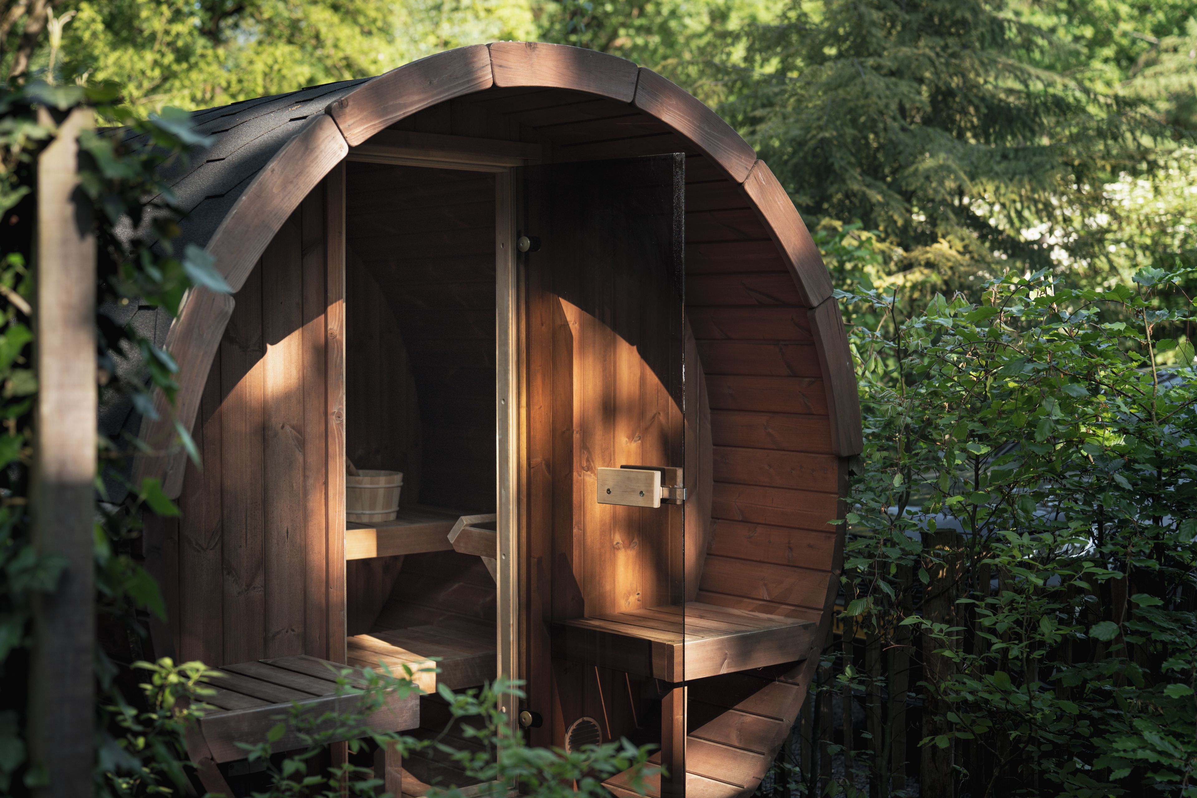Eine Holzsauna mit Sitzbank und Tür, umgeben von viel Grün.