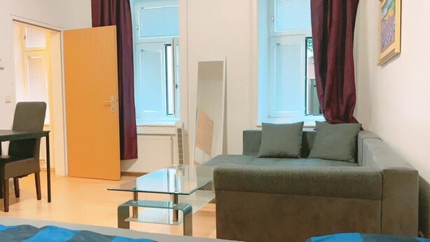 En moderne stue med sofa, sofabord i glas og en lænestol. To vinduer med gardiner, en dør og en radiator.