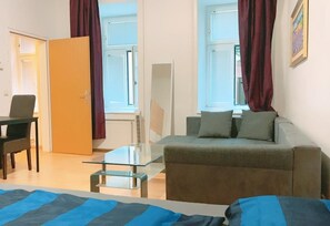 Ein modernes Wohnzimmer mit einer Couch, einem Glastisch und einem Sessel. Zwei Fenster mit Vorhängen, eine Tür und eine Heizung sind vorhanden.