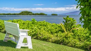 A white bench by a lake with lush greenery and distant hills. - Ocean Views W/Ground Lanai and Kitchen-Molokai Shores (Kaunakakai)