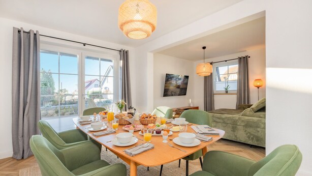 A dining area with a wooden table set for breakfast, green upholstered chairs, and a large window with curtains. - Villa Alma in Loddin - with balcony and terrace and charging station for e-bikes (Loddin)