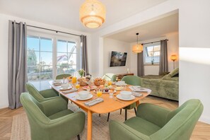 A dining area with a wooden table set for breakfast, green upholstered chairs, and a large window with curtains. - Villa Alma in Loddin - with balcony and terrace and charging station for e-bikes (Loddin)
