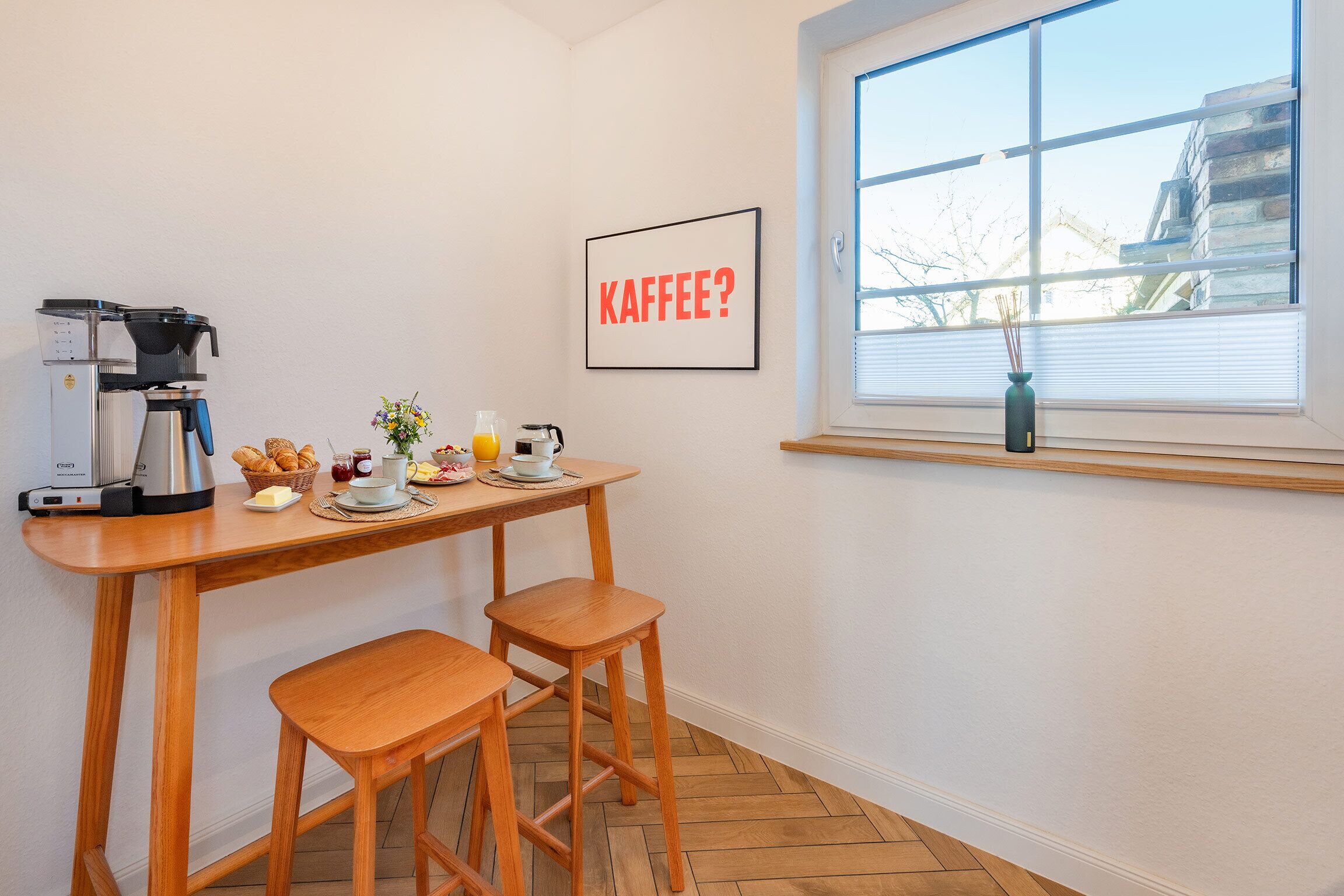 Une petite cuisine équipée d’une machine à café, d’une pancarte encadrée sur laquelle on peut lire « KAFFEE ? », et d’une fenêtre donnant sur les bâtiments environnants.