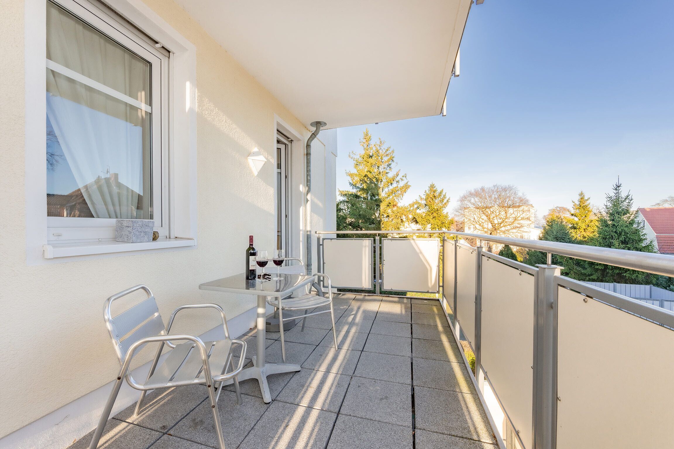 A balcony with a table and chairs, a window with curtains, and a view of trees and buildings.