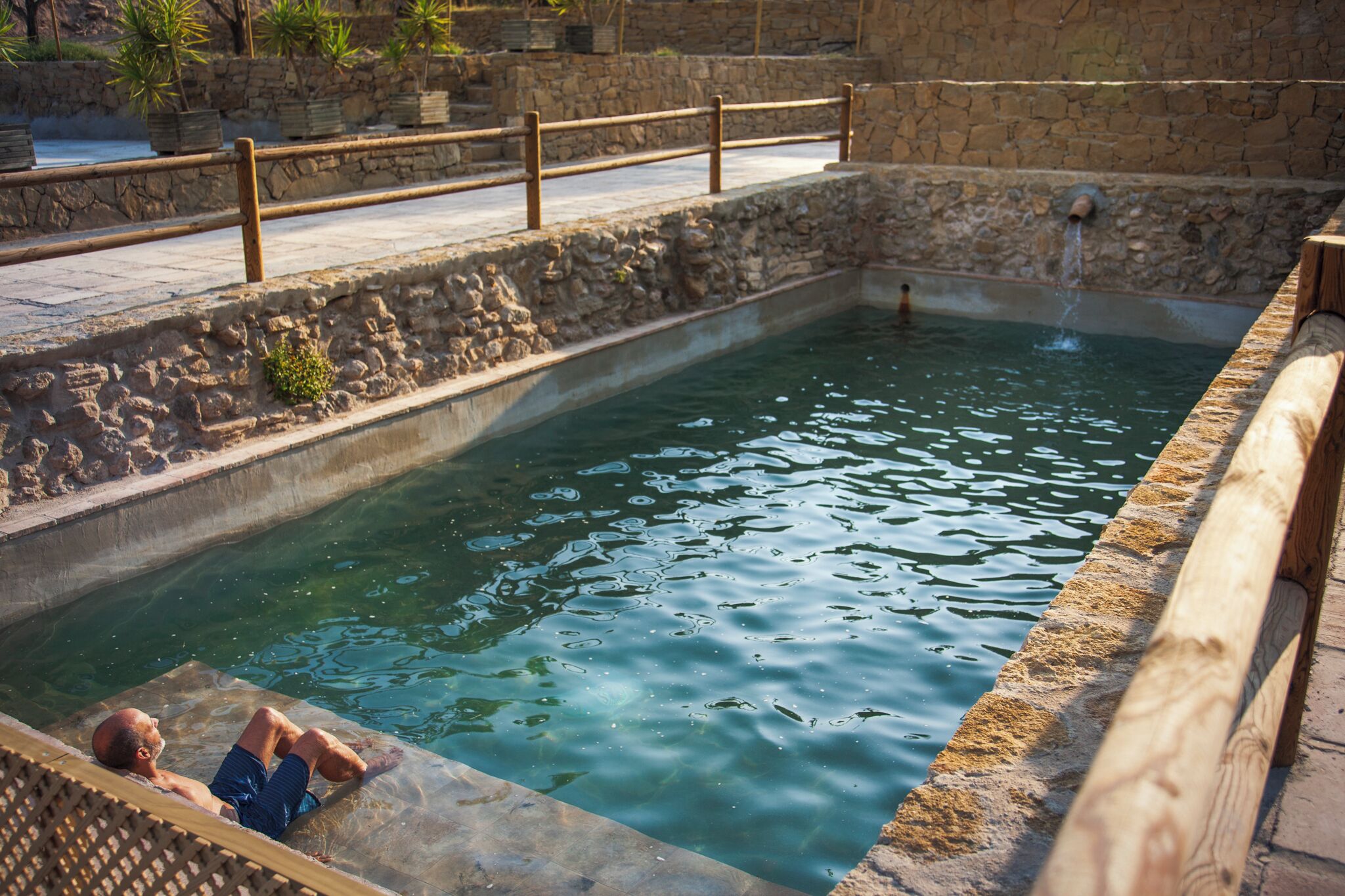 Una piscina de piedra con un muelle de madera y una persona relajándose en el agua.