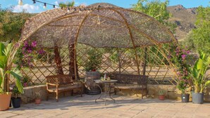A covered outdoor seating area with a wooden bench, a table, and potted plants. - Oasis Al Hamam Wellnesscamp  (Lucainena de las Torres)