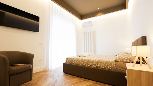 A modern bedroom with a large bed, a sofa, a bedside table, and a flat-screen TV mounted on the wall. - VivimiHome - MaisonErcolano (Ercolano)