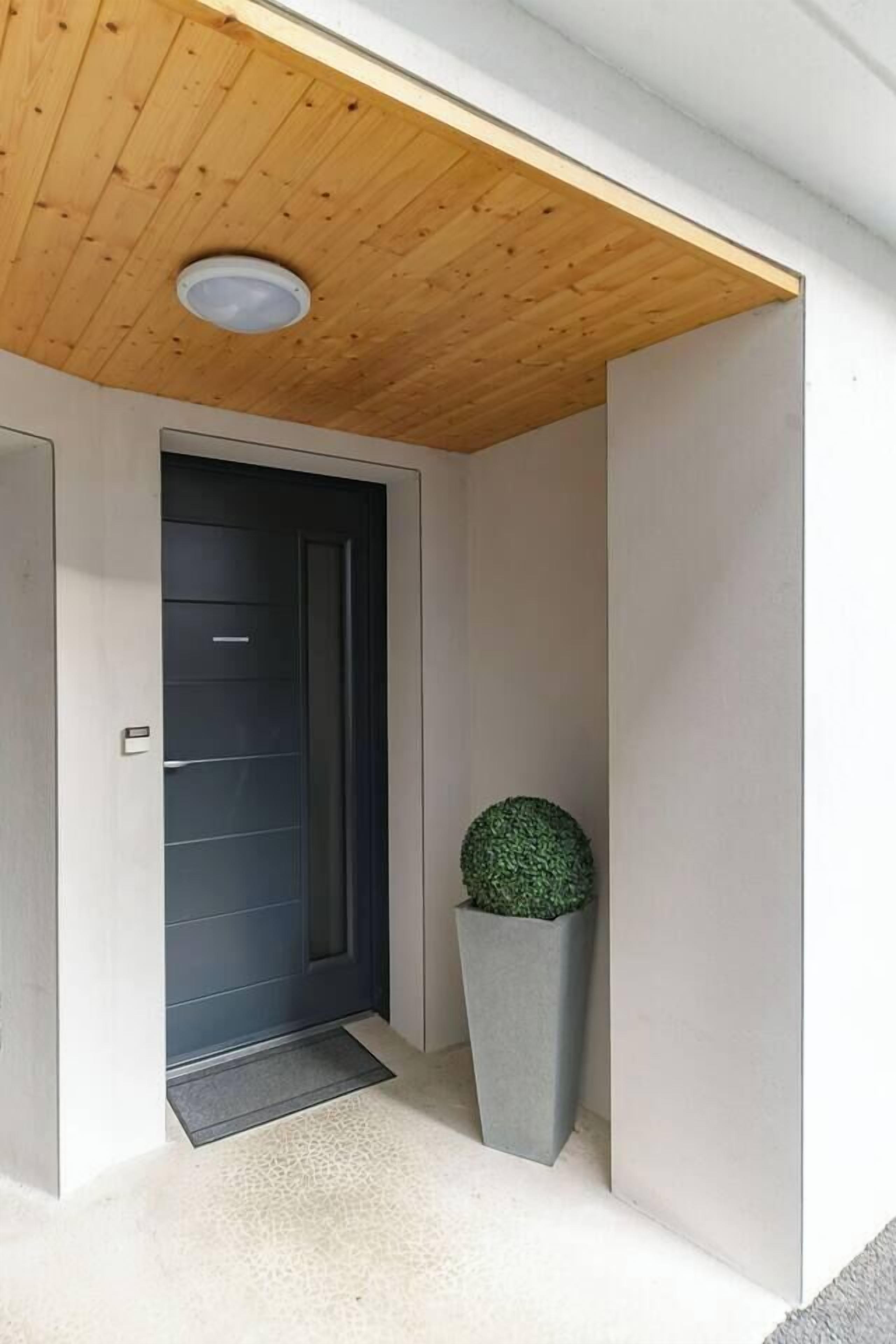A modern entrance with a wooden ceiling, a dark door, and a grey planter with a spherical plant.