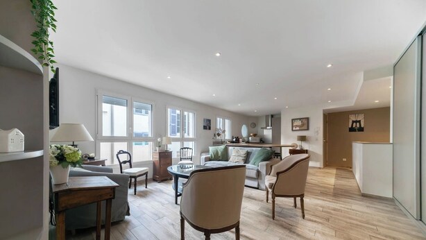 A spacious living area with a wooden floor, a dining table, and several chairs. There is a sofa and a small table with a lamp. The room has large windows and a view of the outside.
