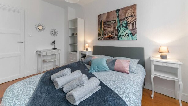 A bedroom with a bed, bedside tables, a desk, a wardrobe, and a wall with a cityscape painting. - Appartement Cosy en centre ville avec parking (Clermont-Ferrand)